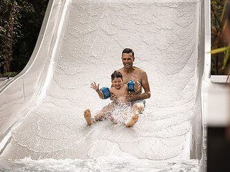 A smiling father and his young son wearing armbands speed down a wide wave water slide together.